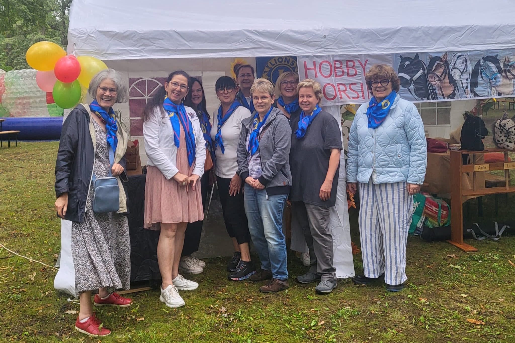 Neun Damen stehen kurz vor Beginn des Müllroser Sommerfests vor dem Aktionszelt mit Hobby Horsing – Steckenpferdparcours und einer Bastelstraße.