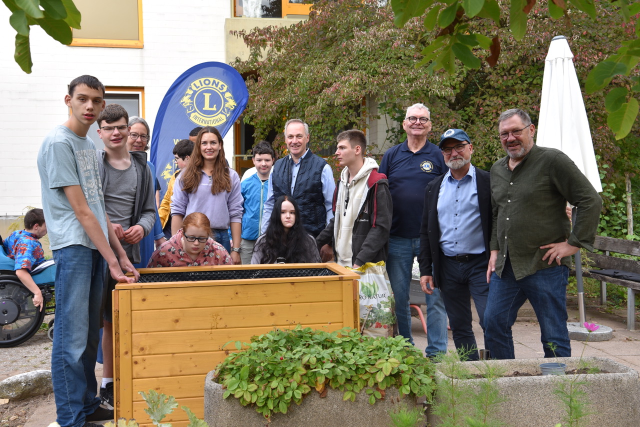 Schüler:innen, Lehrer:innen und Lionsfreunde weihen gemeinsam das 1. Hochbeet ein. Zwei junge Rollstuhlfahrerinnen sitzen am Hochbeet und demonstrieren, dass auch sie das Pflanzen übernehmen könnten.