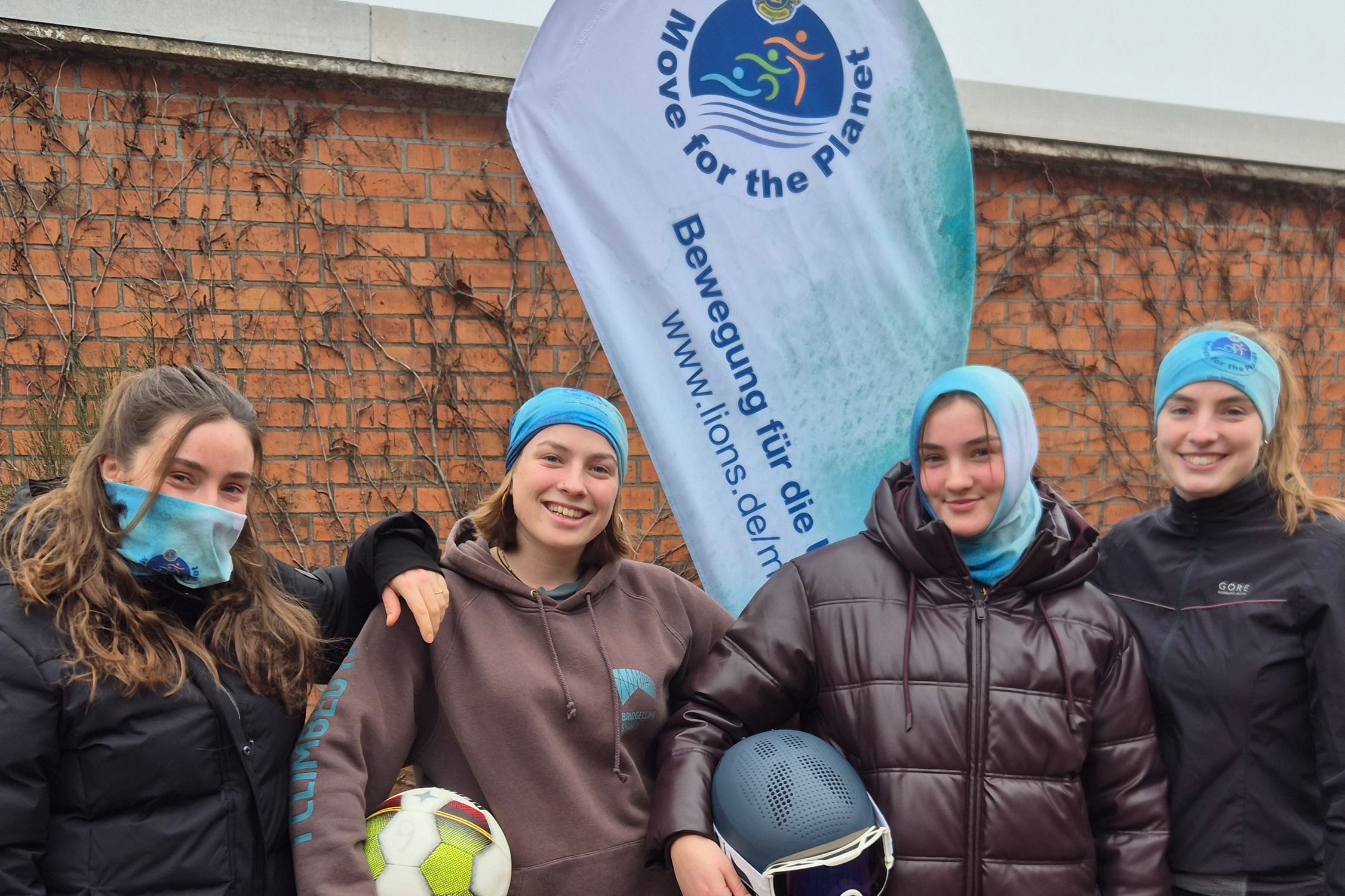 Vier junge Frauen präsentieren vor der Move-Beachflag den Move Loop. Von links nach rechts wird der Loop als Windschutz vor Mund und Nase, als Kopftuch, als Kopfhaube und als Haarband getragen.