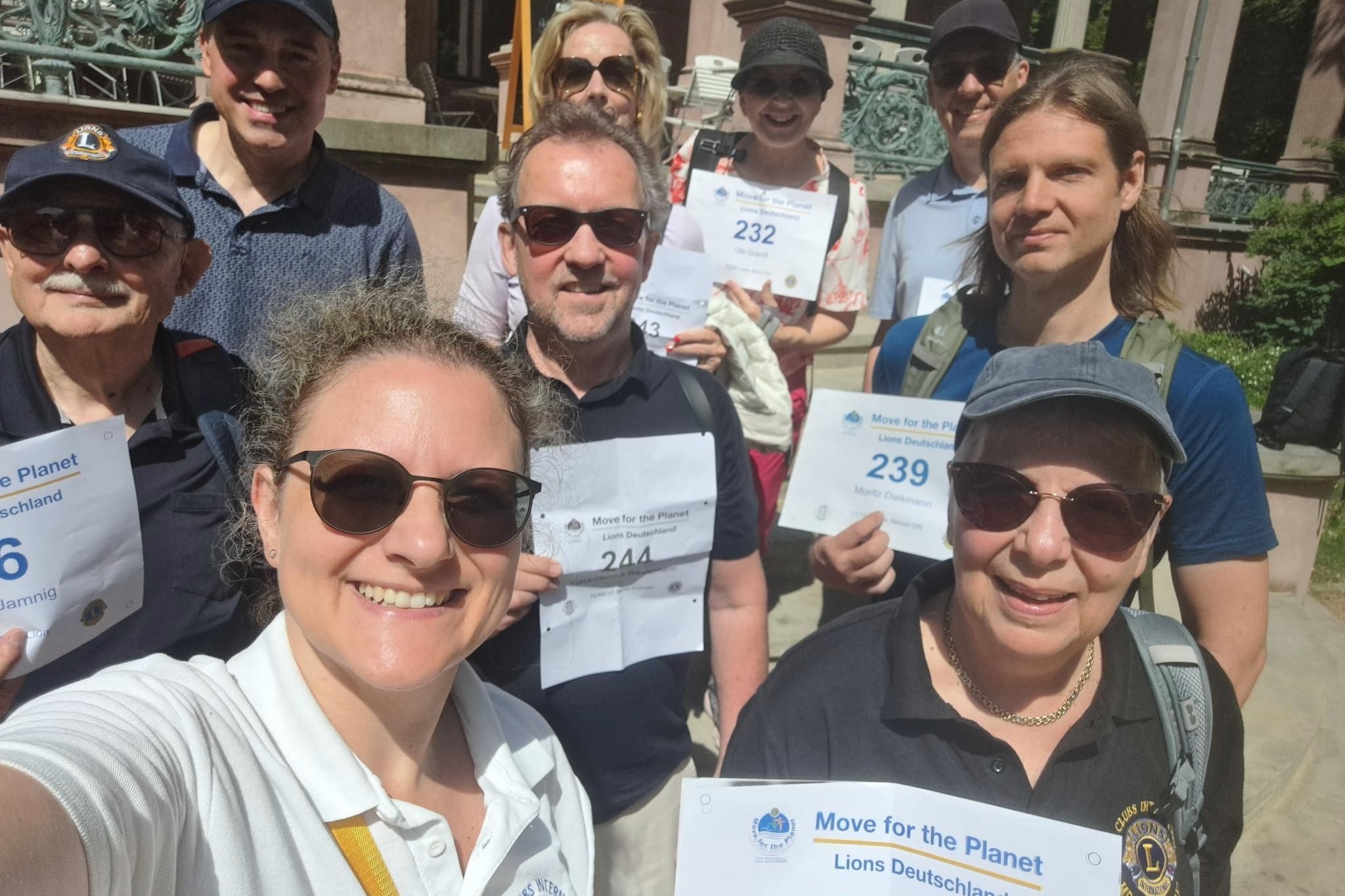 Selfi einer neunköpfigen Gruppe von Erwachsenen, vier Frauen und fünf Männer, die bei einer Stadtwanderung ihre Startnummern hochhalten. Hinter der Gruppe ist eine Hausfassade zu sehen.