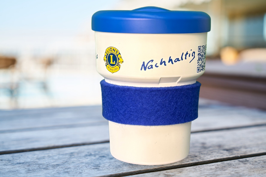 Der LIONS Umweltbecher: Ein weißer Becher mit aufgedrucktem Lions-Emblem, mit blauem Deckel und blauer Filz-Manschette. Der Becher steht auf einem Tisch im Freien.