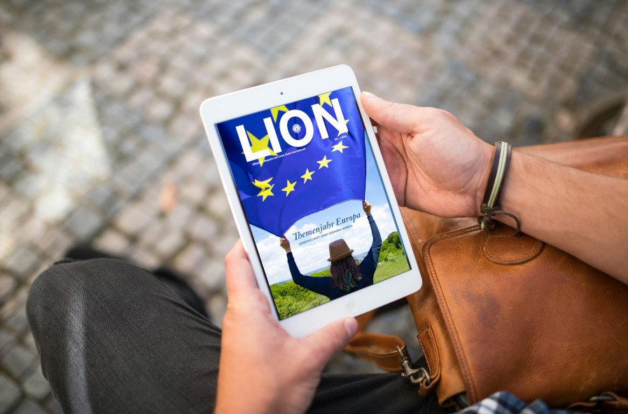 Die aktuelle Ausgabe des Mitgliedermagazins LION wird auf einem Tablet angezeigt. Auf dem Cover ist eine junge Frau in Rückenansicht abgebildet, die auf eine weite Landschaft blickt und eine Europa-Fahne in beiden Händen hält.