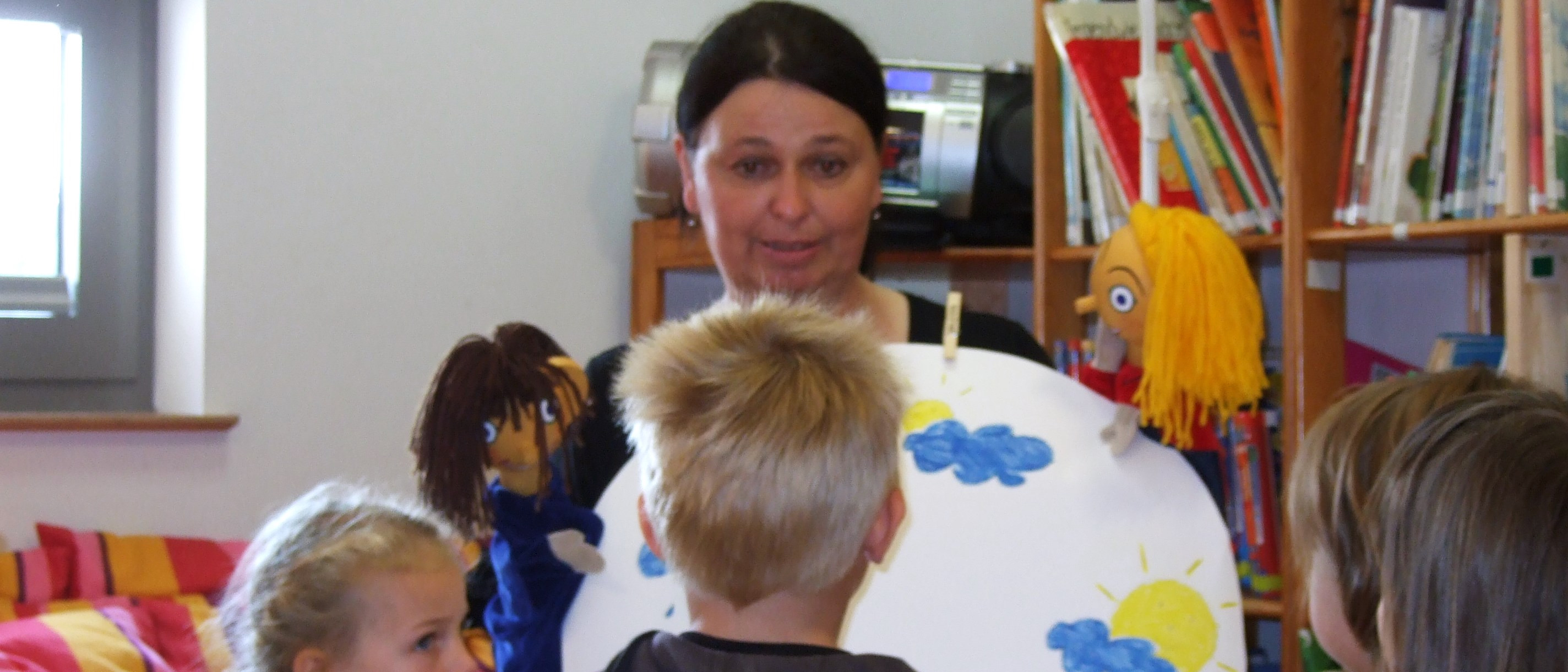 Eine Gruppe Kindergartenkinder hocken vor der Erzieherin, die eine große Scheibe mit Wettersymbolen hochhält und die Kinder bittet zu zeigen, wie sie sich fühlen