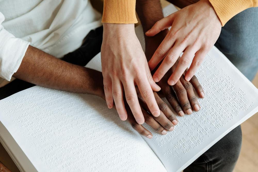 Das Bild zeigt ein aufgeschlagenes Buch mit Brailleschrift, ein paar Hände liest die Brailleschrift gerade, während das andere Paar Hände, das erste über das Papier führt.